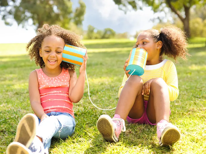 Two young girls sitting on the grass talking to each other through a tin-can telephone