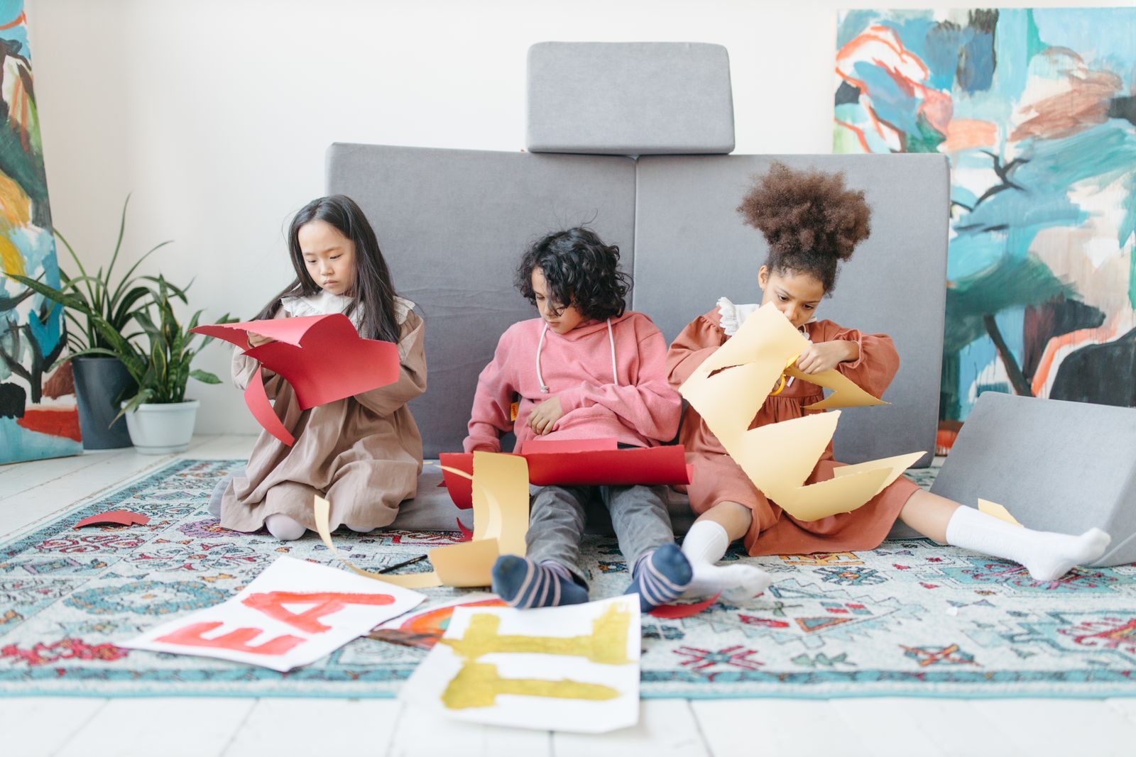 Three children cutting and folding colored paper on a playroom rug